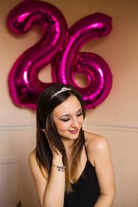 Smiling young woman against number 26 balloon on wall during birthday