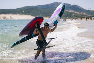 Young shirtless man carrying wing foil at beach