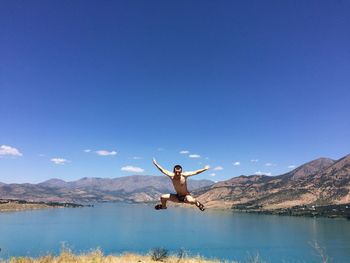 Shirtless man jumping at lake against sky