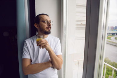 Young man with a beard in a t-shirt and trousers barefoot stands with orange juice at the window