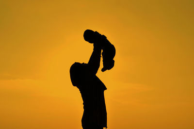 Low angle view of silhouette woman holding aloft baby against orange sky