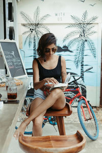 Resting woman in casual wear and trendy sunglasses reading book during refreshment in outdoors bar