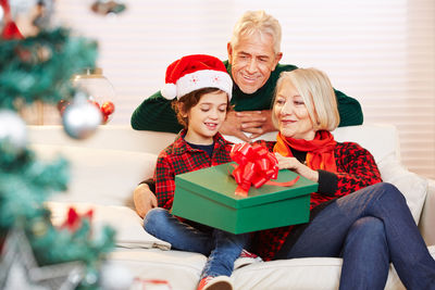 Grandson sitting with grandparents on sofa by christmas tree at home