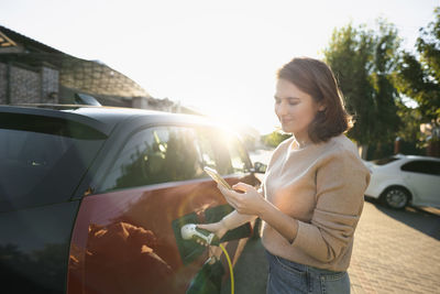 Smiling woman using smart phone and charging car on sunny day