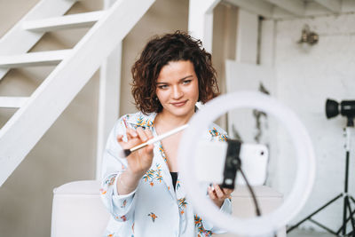 Young woman blogger in home clothes doing makeup with mobile phone and ring lamp online in studio