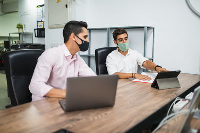 Side view of anonymous entrepreneurs in formal apparel and face masks working on project while surfing internet on netbook at office table