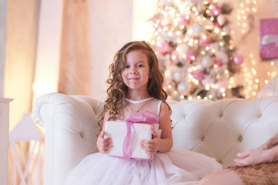 A beatiful girl with curly hair in white dress is holding of christmas box gift and sitting 