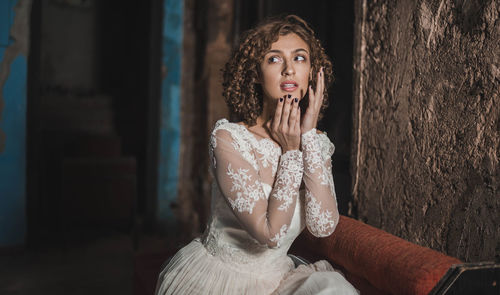 Thoughtful woman in wedding dress sitting with hand on chin at home
