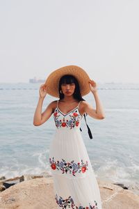 Portrait of young woman wearing hat standing at beach against sky