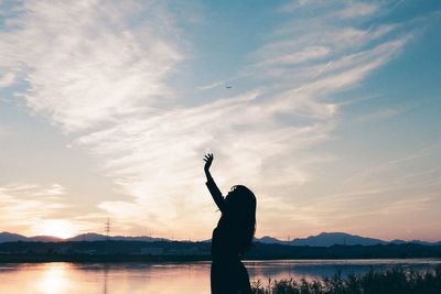 Silhouette woman with hand raised standing by lake against sky during sunset