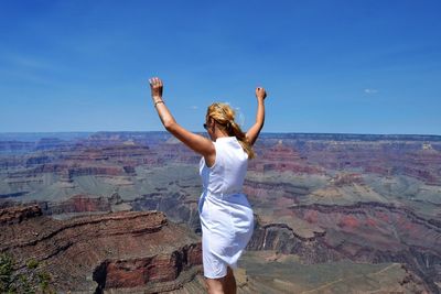 Rear view of woman with arms raised standing by grand canyon national park against blue sky