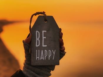 Cropped hand of woman holding be happy text on tag against sea during sunset