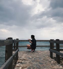 Woman crouching on bridge over sea against sky