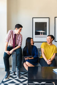 Smiling male and female business colleagues talking while sitting on sofa against wall at office