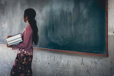 Side view of woman carrying books while walking against blackboard in classroom