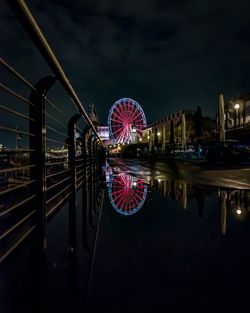 Illuminated ferris wheel by river against sky at night