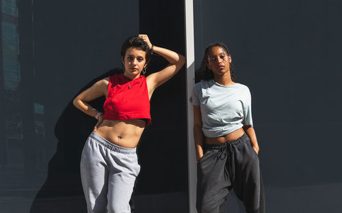 Serious multiracial female friends in casual clothes looking at camera while standing near gray wall of building on sunny street