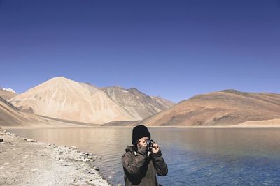 Man photographing at lakeshore against clear blue sky