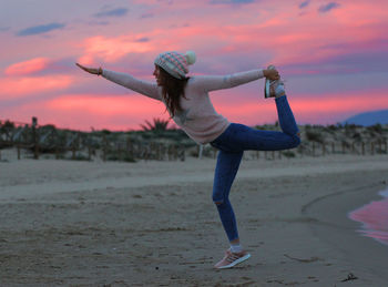 Woman doing yoga at beach against sky during sunset