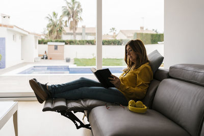 Relaxed woman reading book while sitting on reclining chair at home