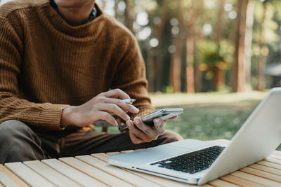 Midsection of man using mobile phone sitting by laptop