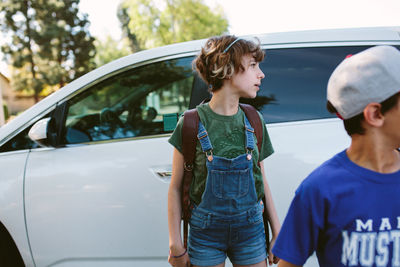 Profile shot of girl in overall shorts and backpack stands next to car