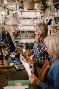 Senior female owner reading documents by coworker talking on mobile phone at checkout in store