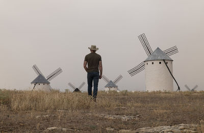 Rear view of man with cowboy hat standing on field with spanish white windmills against sky