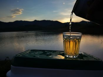 Close-up of glass on table against lake during sunset