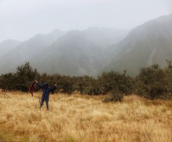 Hooded person holding red fabric on grassy field by mountains against sky