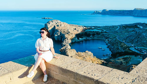A beautiful caucasian brunette sits on the curb of the acropolis against the backdrop of a seascape.