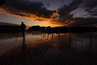 Silhouette person standing on footbridge against sky during sunset