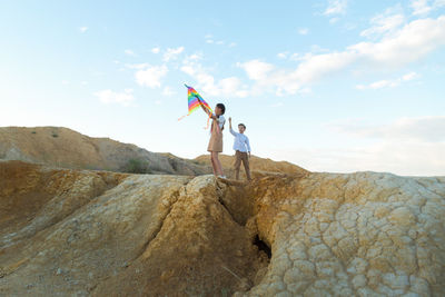 Brother and sister launch bright big kite into sky near a flowing river.