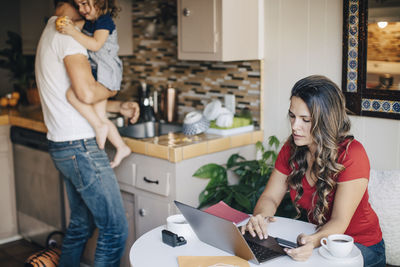 Mother using laptop while father carrying daughter and working in domestic kitchen