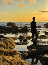 Man standing on rock by sea against sky during sunset