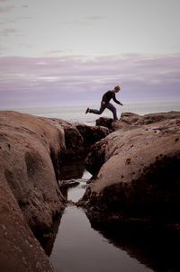 Side view of man jumping on rock at beach against sky during sunset