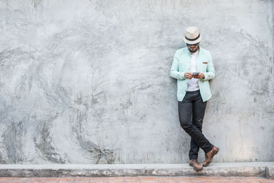 Young man using phone while standing against concrete wall