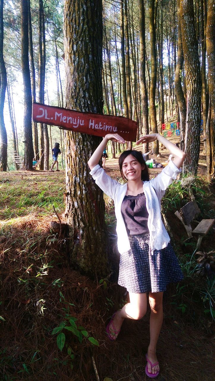 tree, real people, one person, young adult, lifestyles, leisure activity, young women, forest, front view, full length, tree trunk, arms raised, smiling, looking at camera, standing, day, happiness, growth, nature, outdoors, portrait, beautiful woman, beauty in nature