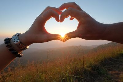 Cropped hands of couple gesturing heart shape against sun during sunset