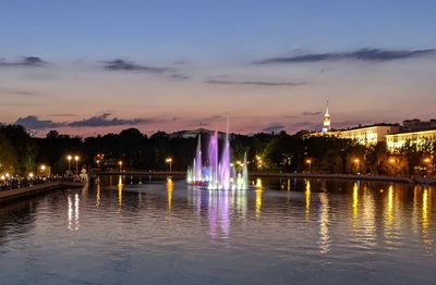 Illuminated buildings by river against sky at sunset