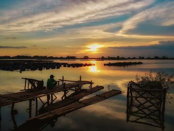 Rear view of silhouette man crouching on pier over lake at sunset