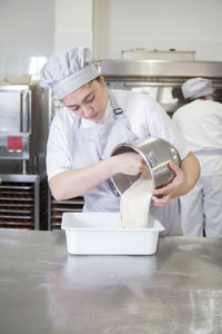 Side view of female baker in uniform putting raw dough from bowl into container while working in professional light bakery