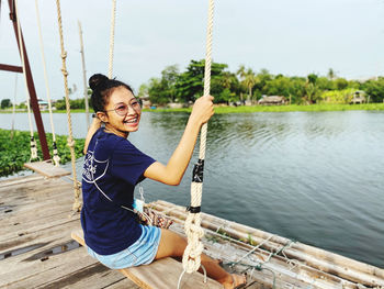 Smiling young woman sitting on rope against lake
