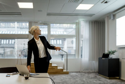 Confident businesswoman using laptop while looking at flat screen in board room