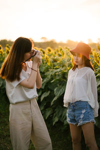 Woman photographing female friend standing at sunflower farm