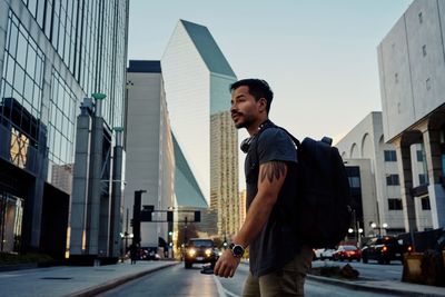 Side view of hispanic male traveler in casual clothing with backpack and headphones on neck walking along modern city street