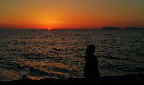 Silhouette boy standing on beach against orange sky