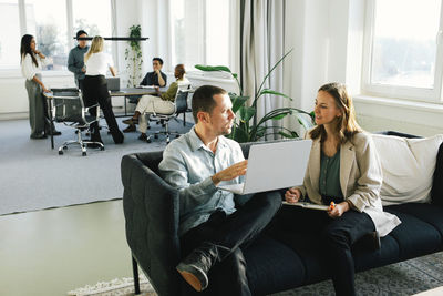 Businessman explaining female colleague over laptop while sitting on sofa at office