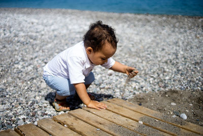 Happy kid in white shirt and blue shorts plays with pebble on the beach