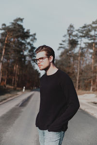 Young man with glasses standing in road against trees and sky